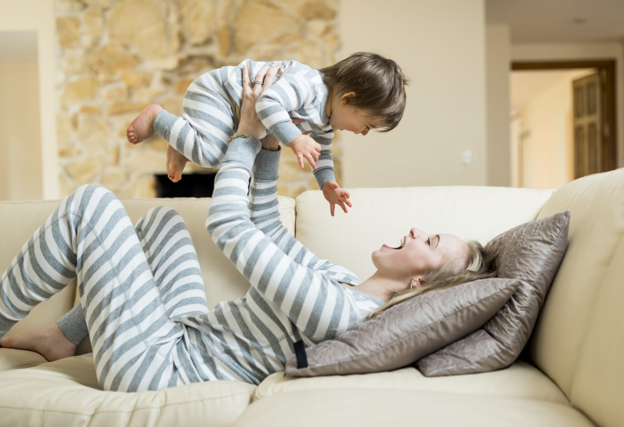 A mother picks up her smiling son above her while reclined on the couch. They are both wearing matching striped pajamas on the couch.