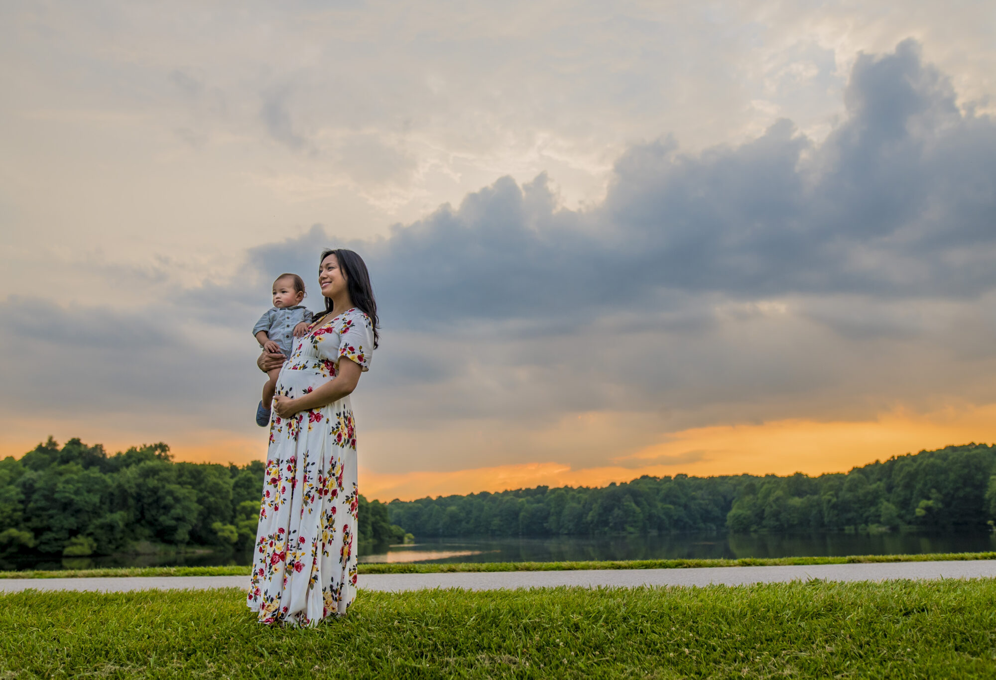 A pregnant mother in a white patterned dress holds her son at sunset with blue clouds and an orange sky behind her.