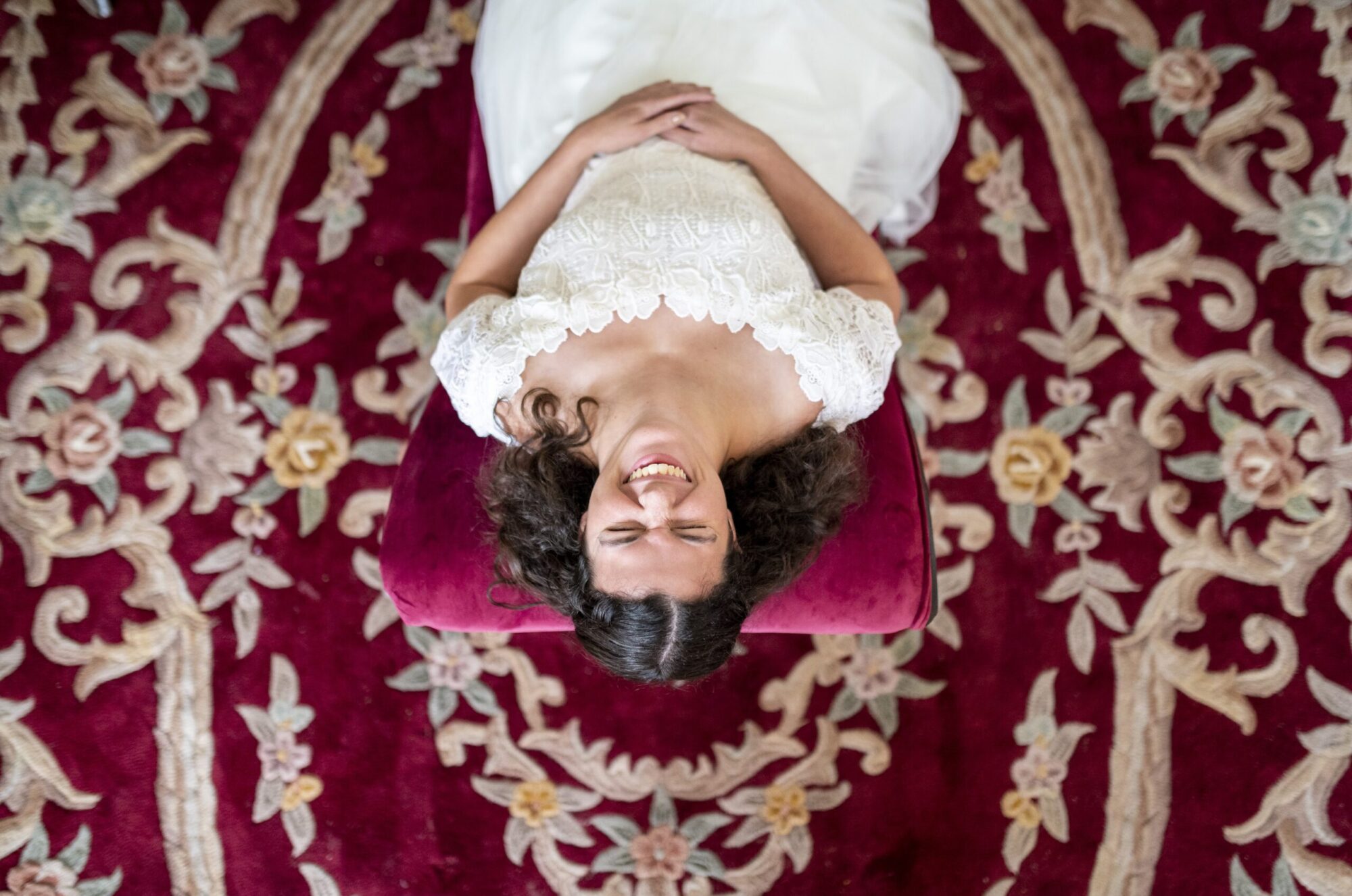 A bride sitting in a chair looking up and smiling and laughing surrounded by red carpet with patterns on her wedding day. Family and wedding photography in Pittsburgh.