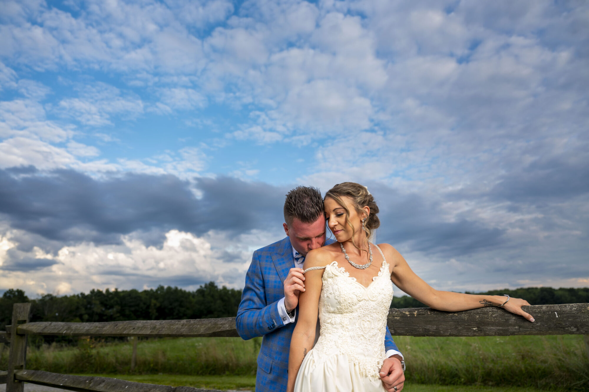 A groom in a blue suit kissing the bare shoulder of his bride in a beautiful landscape with cloudy blue sky. Bride wears a dress with a shoulder strap being pulled down by the groom. Family and wedding photography in Pittsburgh.